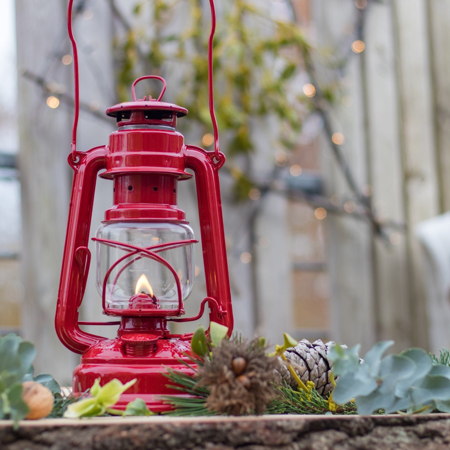 Handcrafted bright red galvanized oil lantern with thermal heat-resistant SUPRAX globe lit outdoors on rustic wooden surface surrounded by seasonal greenery, ideal for Lamps & Lighting, Camping, Emergency Supplies, and Outdoors collections.
