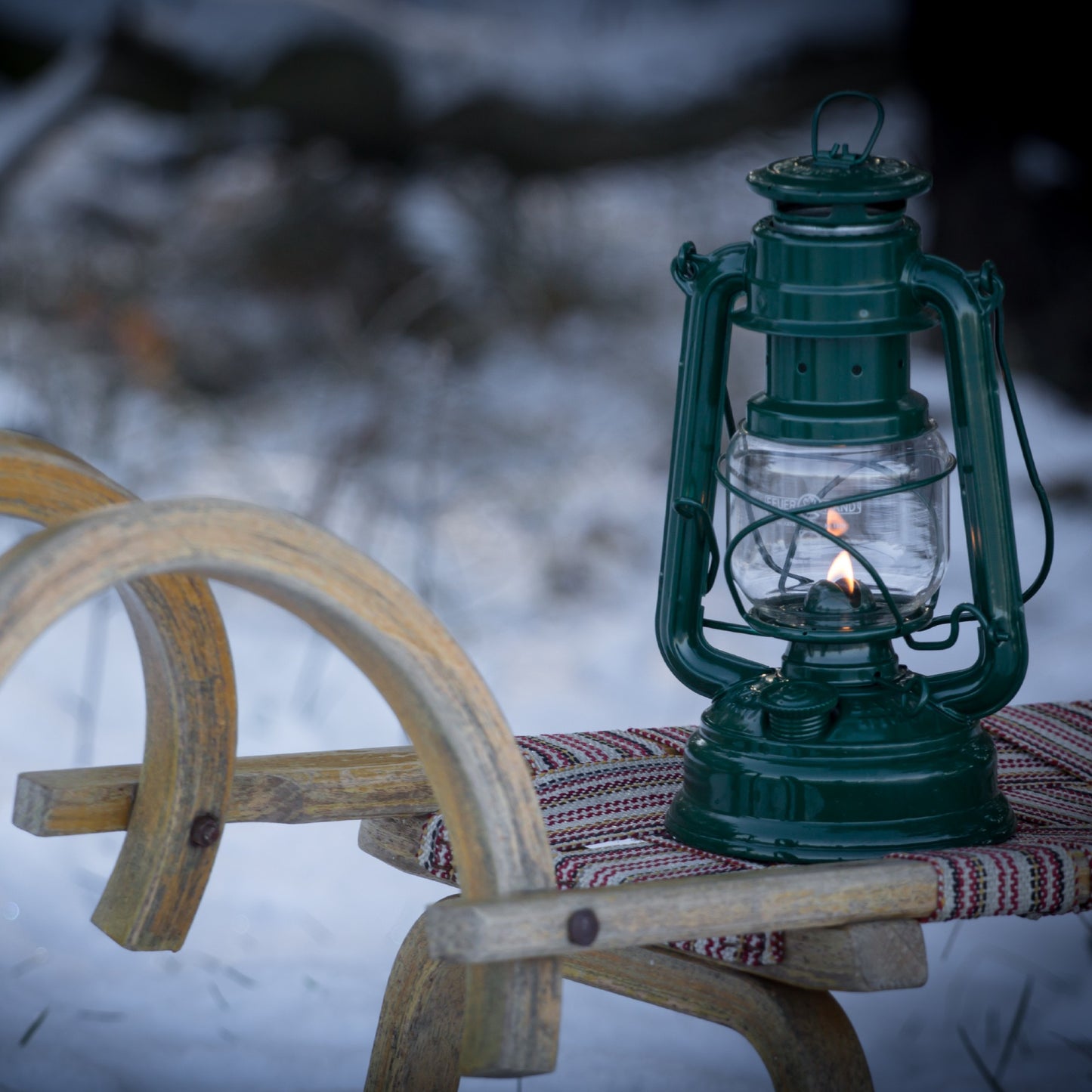Handcrafted green galvanized oil lantern with thermal heat-resistant SUPRAX globe, glowing gently on a rustic wooden sled in a snowy outdoor setting, ideal for camping, emergency supplies, and lamps & lighting collections.
