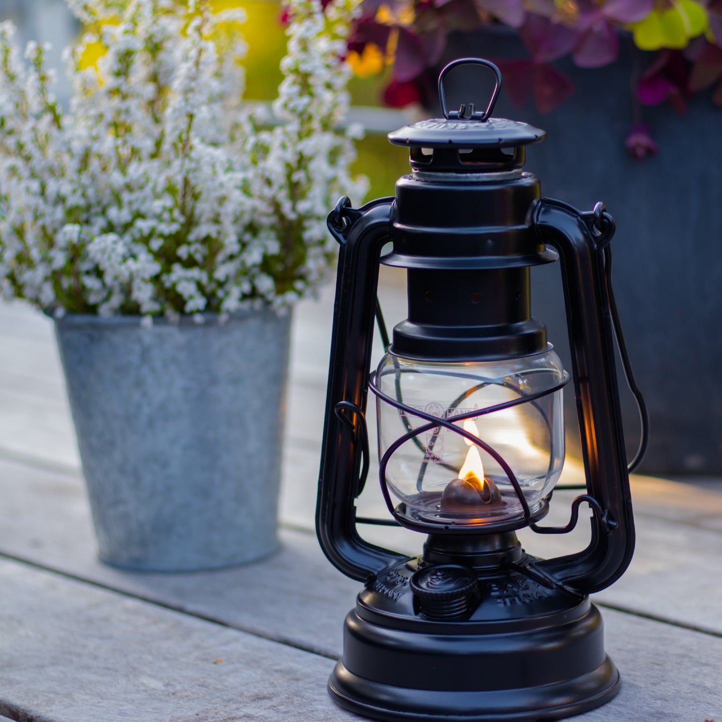 Handcrafted black galvanized oil lantern with thermal heat-resistant SUPRAX globe, glowing softly on a wooden surface near potted flowers, ideal for Lamps & Lighting, Camping, and Emergency Supplies.
