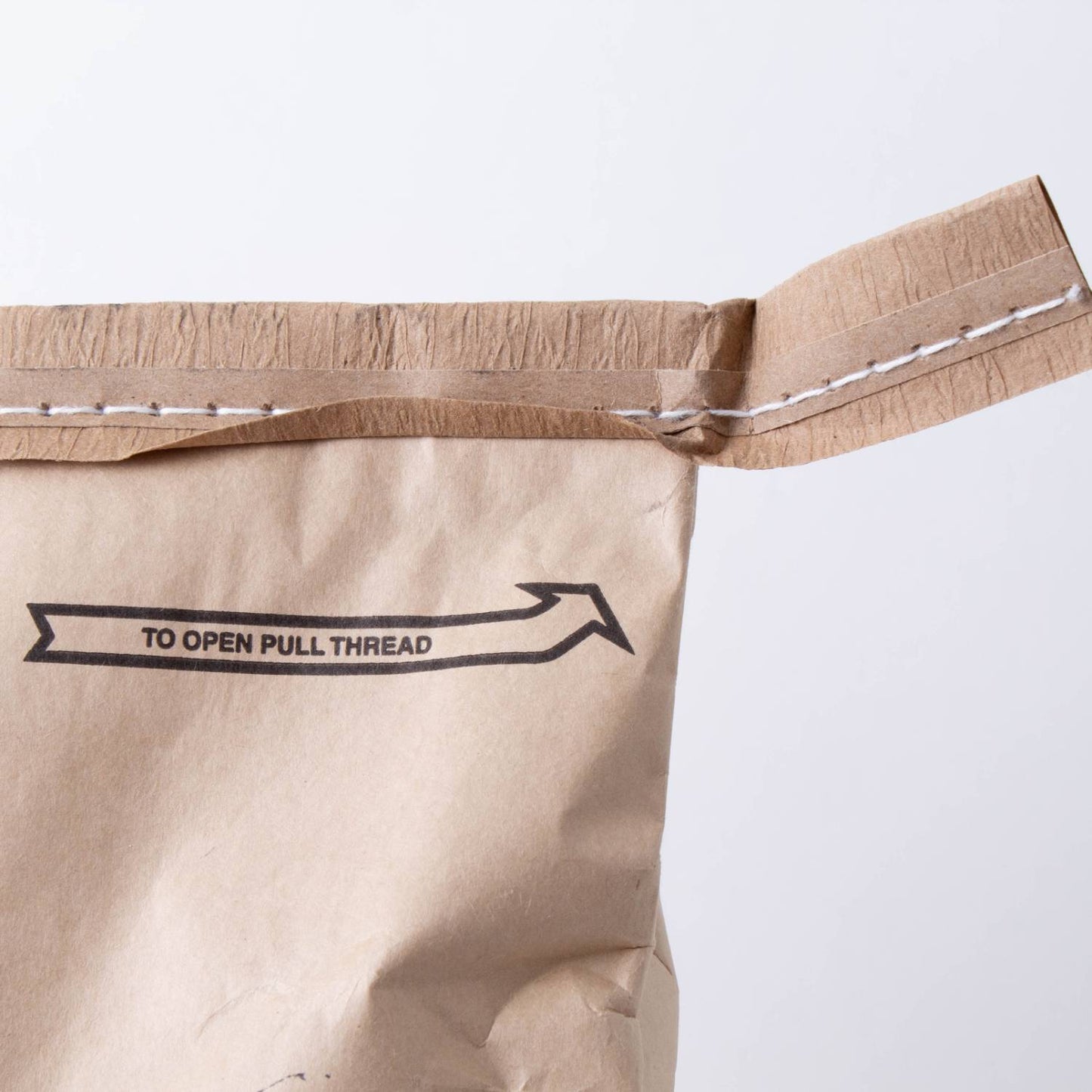 Close-up of a sealed brown paper bag with a stitched pull thread and printed arrow indicating how to open, suitable for pantry, food storage, and emergency supplies collections.