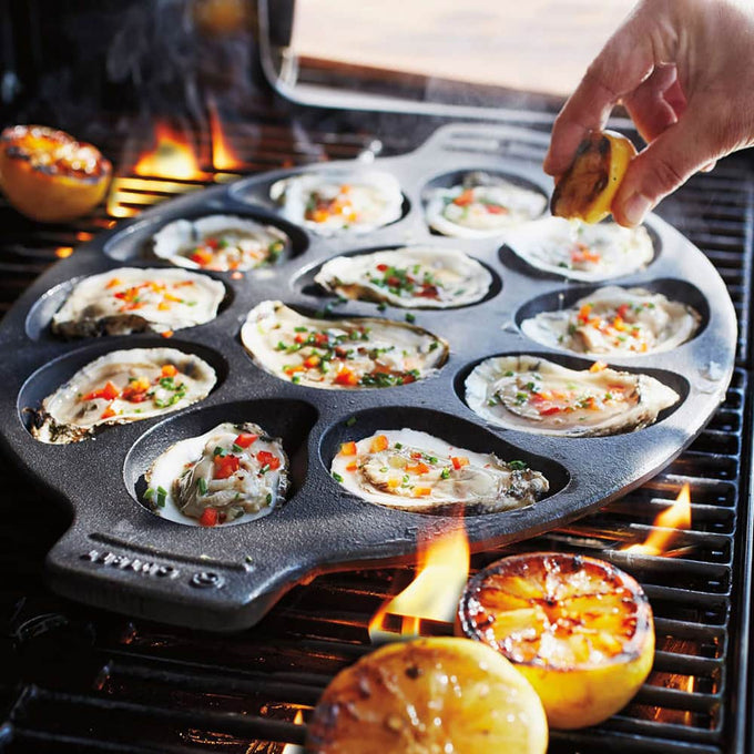 Oval cast iron pan with a dozen oysters grilling on a barbecue, garnished with diced peppers, beside grilled lemon halves, showcasing durable cookware for Kitchen & Food and Grilling collections.