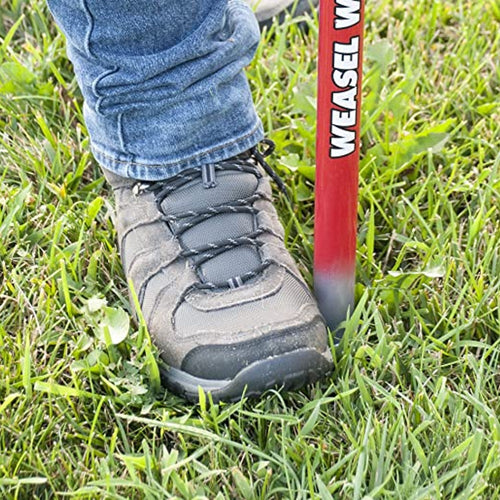 Close-up of a person wearing sturdy shoes pressing the red steel shaft of a handcrafted Garden Weasel WeedPopper into green grass, part of Farm, Garden & Tool and Digging and Cultivating collections.