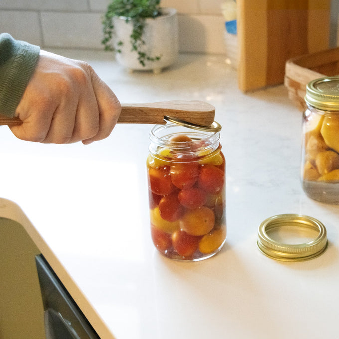 Handcrafted wooden bubble popper tool used to release air bubbles from a jar filled with colorful preserved tomatoes, set in a bright kitchen for food preservation collections including Kitchen & Food and Canning Accessories.