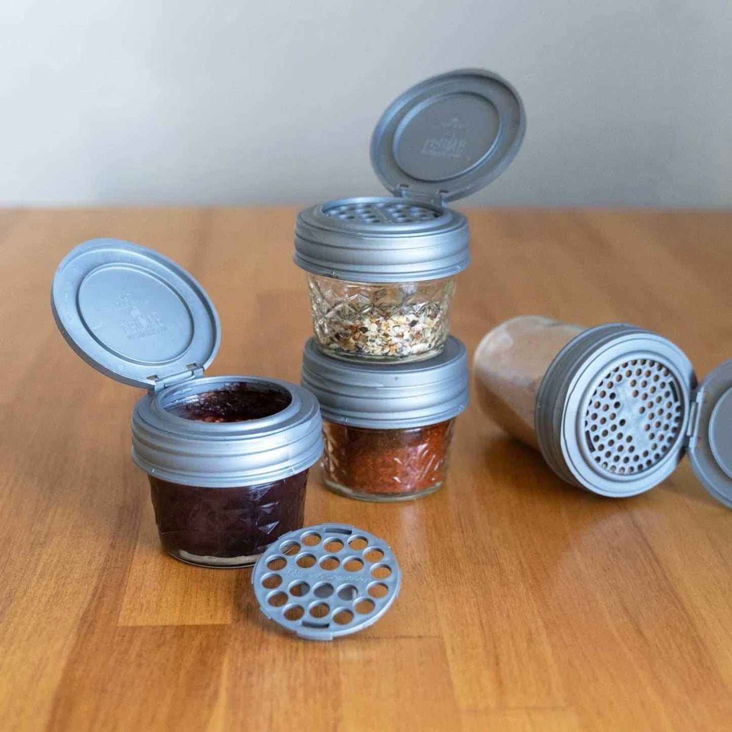 Four silver airtight shaker lids with flip tops and interchangeable inserts, fitted on regular mouth jars containing spices and seasonings, featured in Kitchen & Food and USA Made collections.
