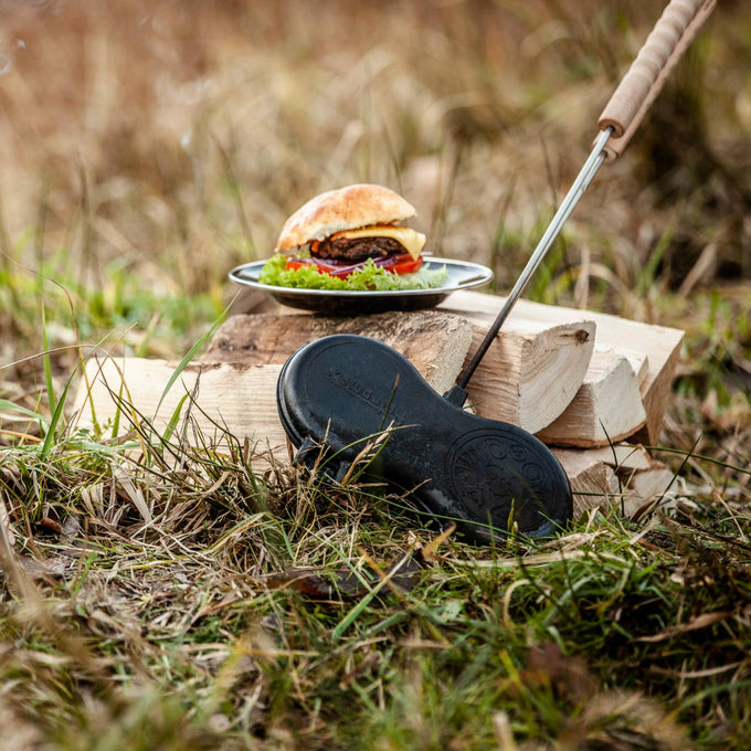 Cast iron burger iron with wooden handle resting outdoors on grass near firewood and a plated burger, designed for camping, outdoor cooking, and cookware collections.