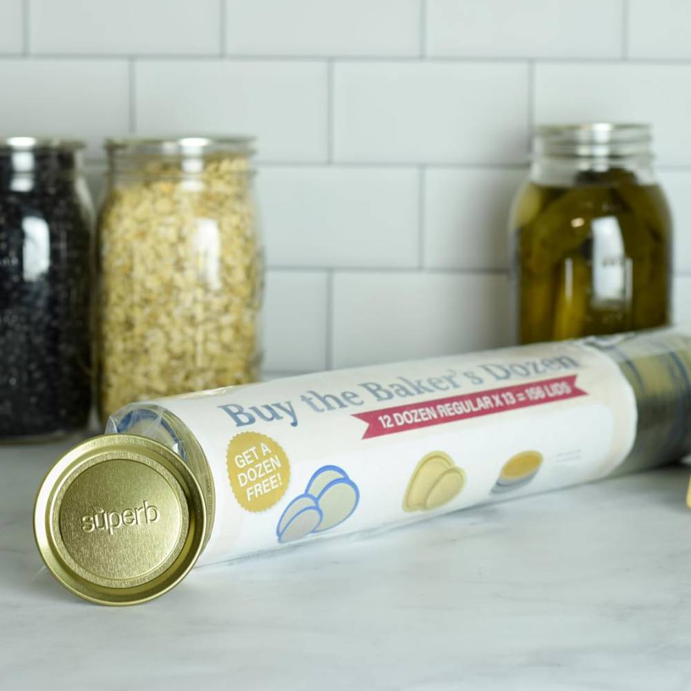 Roll of gold-tone metal regular-mouth canning jar lids embossed with "superb," displayed on a kitchen countertop with jars of preserved food in the background, part of USA made food preservation collections.