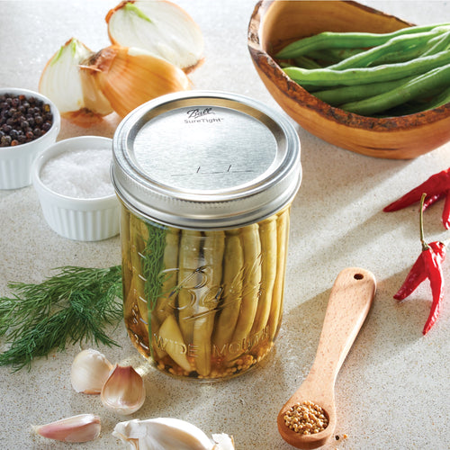 Clear wide-mouth pint jar filled with pickled green beans, sealed with metal lid, surrounded by fresh garlic, dill, onions, spices, and green beans in a wooden bowl, showcasing handcrafted, made in USA canning jar ideal for Kitchen & Food, Food Preservation, and Canning collections.