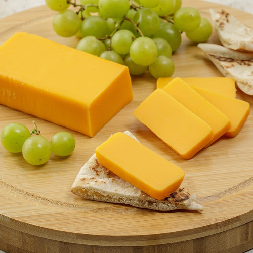 Block of handcrafted shelf-stable cheese sliced and placed on a piece of flatbread, accompanied by green grapes on a round wooden board, ideal for Kitchen & Food, Pantry, and USA Made collections.