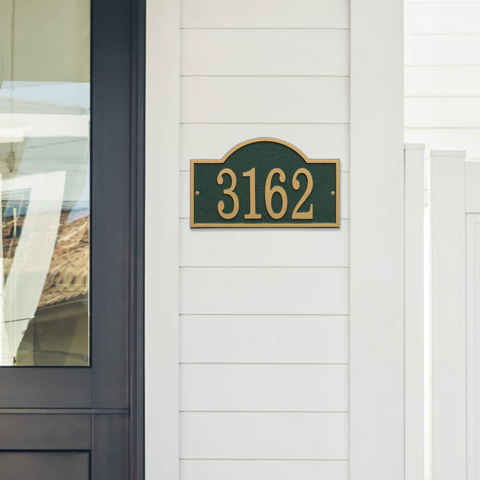 Handcrafted green and gold arched address plaque with bold numbers mounted on white siding, designed to enhance curb appeal, made in USA, ideal for Home & Decor and Mailboxes and Address Signs collections.