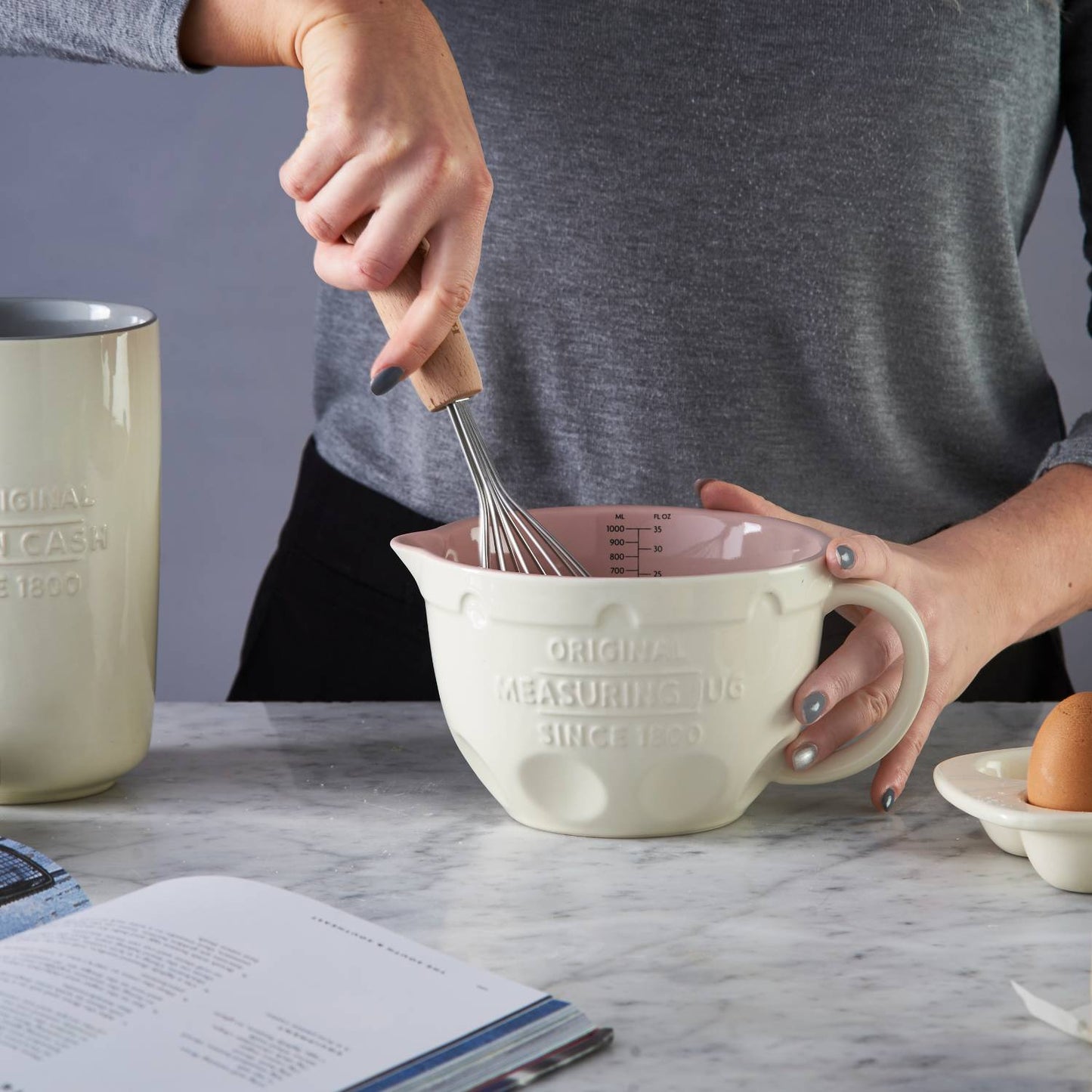 Person whisking batter or mixture in a cream-colored Mason Cash measuring jug with pink interior on marble countertop alongside egg and cookbook, featured in Kitchen & Food, Cookware, Baking Supplies, Food Prep, Measuring, Best Sellers, Promo Eligible collections