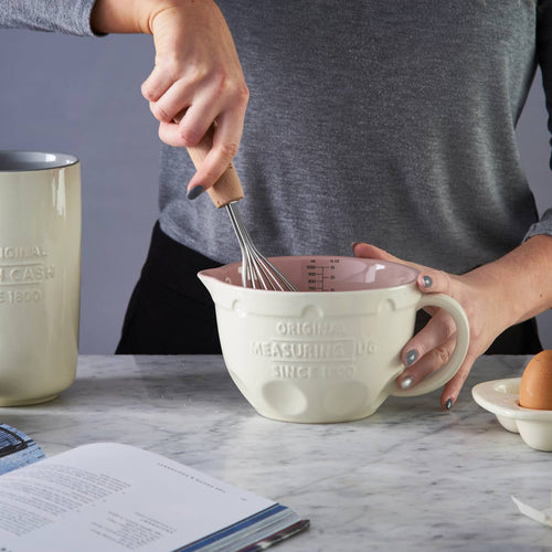 Person whisking batter or mixture in a cream-colored Mason Cash measuring jug with pink interior on marble countertop alongside egg and cookbook, featured in Kitchen & Food, Cookware, Baking Supplies, Food Prep, Measuring, Best Sellers, Promo Eligible collections