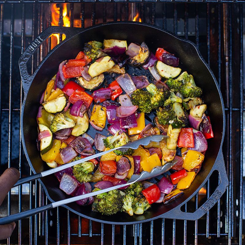 Handcrafted cast iron grilling basket with dual handles, filled with grilled colorful vegetables including broccoli, red and yellow peppers, and onions, cooking over open flame, ideal for Kitchen & Food, Cookware, Cast Iron, Grilling, and Best Sellers collections.