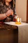 Person holding a spoon over a small glass jar oil lamp with a burning flame inside, set on a cloth-covered table next to a lighter, in a warm indoor setting, showcasing handcrafted USA made lamps & lighting parts.