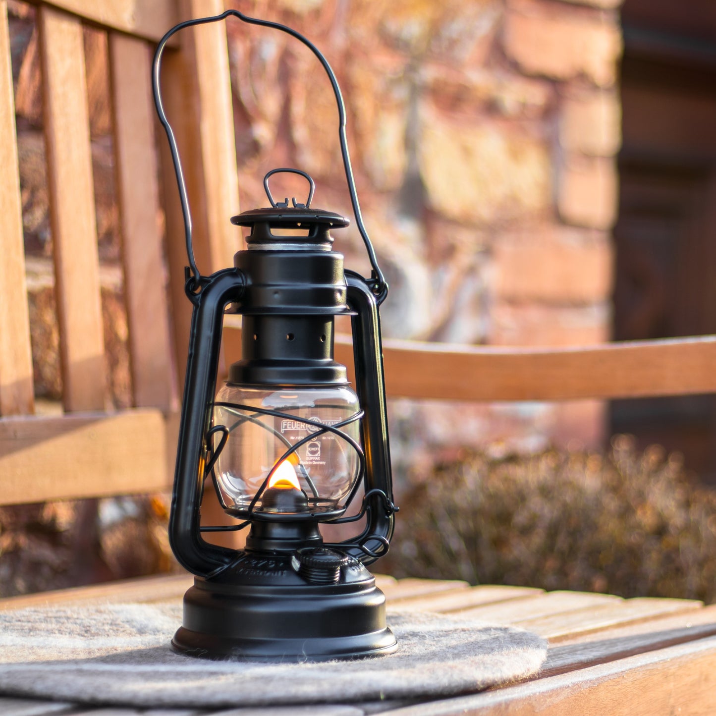 Handcrafted black galvanized oil lantern with thermal heat-resistant SUPRAX globe, softly glowing on a wooden bench outdoors, ideal for camping, emergency supplies, and lamps & lighting collections.
