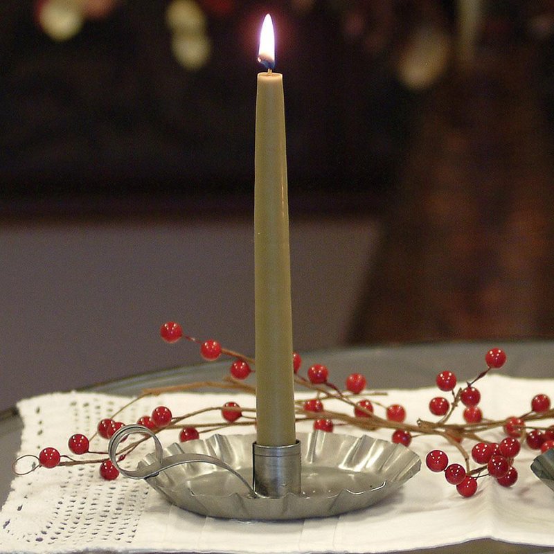 Handcrafted olive-green bayberry candle burning in a rustic pewter holder, displayed with decorative red berries on a white lace cloth, reflecting traditional holiday decor.