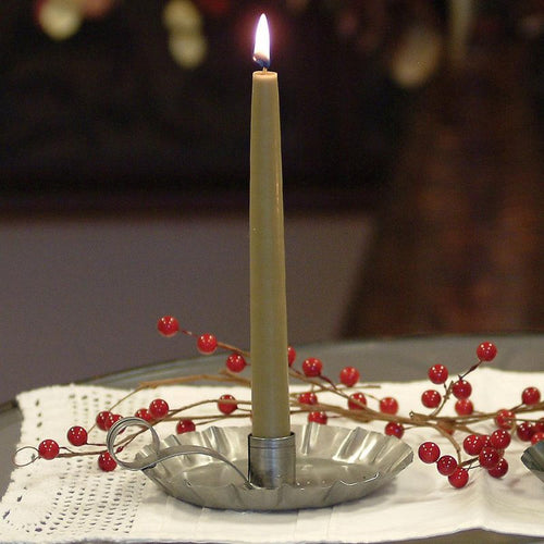 Handcrafted olive-green bayberry candle burning in a rustic pewter holder, displayed with decorative red berries on a white lace cloth, reflecting traditional holiday decor.