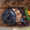 Seasoned cast iron smoker skillet partially covered with a vented lid, filled with wood smoking chips on a wooden cutting board, alongside fresh rosemary and orange peel, featured in Kitchen & Food, Cookware, Cast Iron, USA Made, Best Sellers, and Promo Eligible collections.