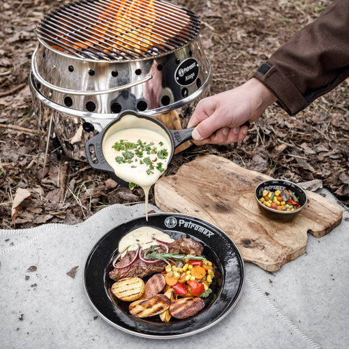 Hand pouring creamy sauce from a small cast iron pan over grilled steak and vegetables on a black plate outdoors, next to a fire pit, suitable for Farm, Garden & Tool, Kitchen & Food, Camping, Cookware, Cast Iron, Best Sellers, and Promo Eligible collections.