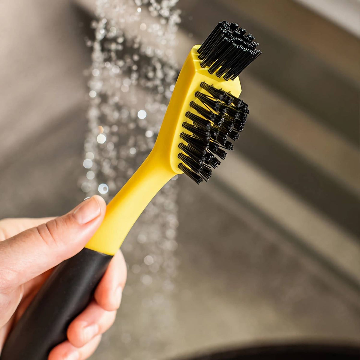 Hand holding a yellow and black cleaning brush with two nylon bristle heads under running water, designed for deep cleaning corners and grooves, ideal for Kitchen & Food, Cookware, Cast Iron, Best Sellers, and Promo Eligible collections.