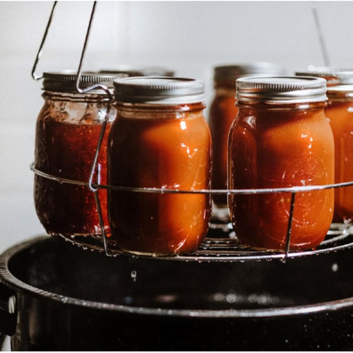 Glass jars filled with homemade preserves in a metal canning rack, being processed in a large black enamelware pot, featured in Kitchen & Food and Food Preservation collections.