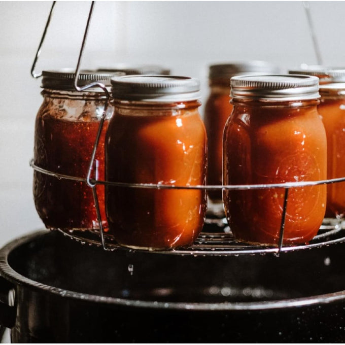 Glass jars filled with homemade preserves in a metal canning rack, being processed in a large black enamelware pot, featured in Kitchen & Food and Food Preservation collections.