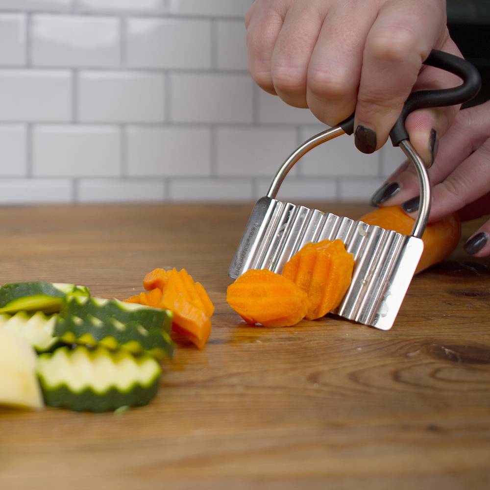 Handheld vintage-style crinkle cutter slicing bright orange carrots into decorative ridged pieces on wood surface, ideal for Kitchen & Food, Food Prep, and Choppers and Cutters collections.