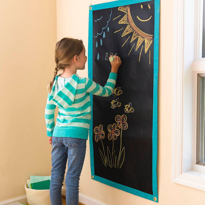 Child drawing colorful flowers and sun with chalk on a portable roll-up chalk mat hanging on a wall, featured in Toys, Books & Games and Children's Toys collections.