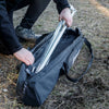 Person placing a metal cooking tripod into a black transport bag with reinforced base outdoors on dry grass, suitable for camping and farm use in Best Sellers and Promo Eligible collections.