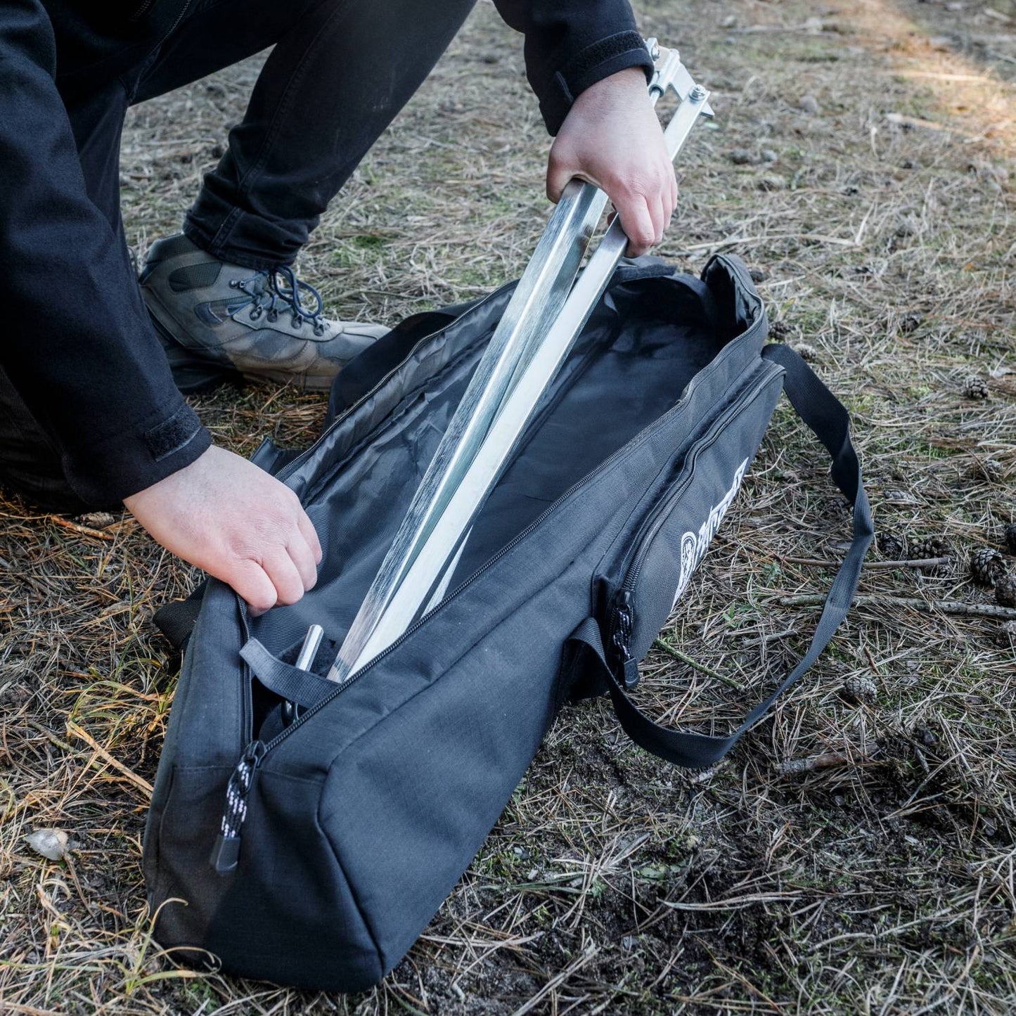 Person placing a metal cooking tripod into a black transport bag with reinforced base outdoors on dry grass, suitable for camping and farm use in Best Sellers and Promo Eligible collections.