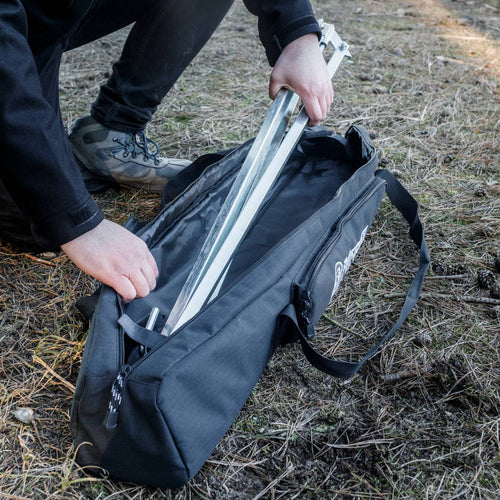Person placing a metal cooking tripod into a black transport bag with reinforced base outdoors on dry grass, suitable for camping and farm use in Best Sellers and Promo Eligible collections.
