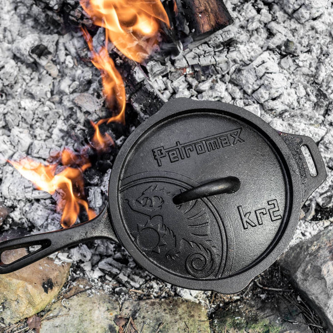 Cast iron saucepan with self-basting lid cooking over hot embers, featuring precision pouring aids, ideal for farm, garden, camping, cookware, and cast iron collections.