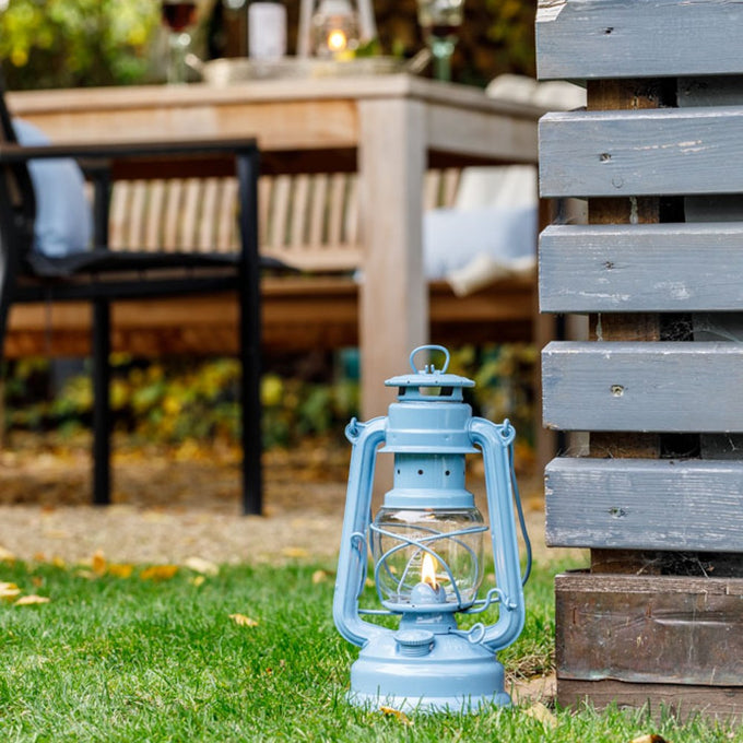 Handcrafted light blue galvanized oil lantern with a glowing thermal heat-resistant SUPRAX globe, placed outdoors near rustic wooden furniture, ideal for camping and emergency supplies collections.
