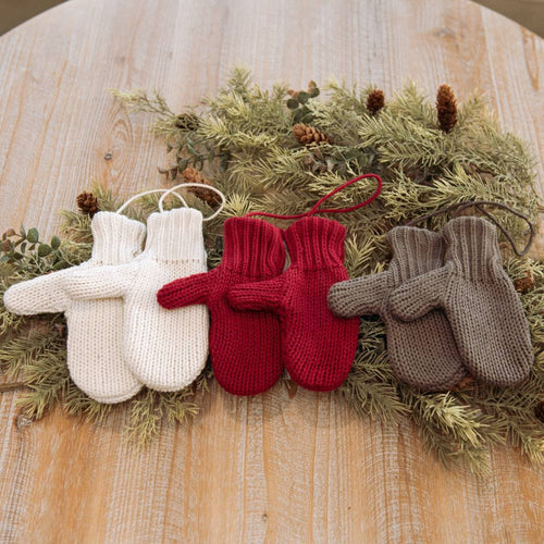 Three pairs of knitted Christmas mitten decorations in cream, red, and brown hanging on natural greenery with pine cones, featured in Home & Decor and Holiday Decor collections.