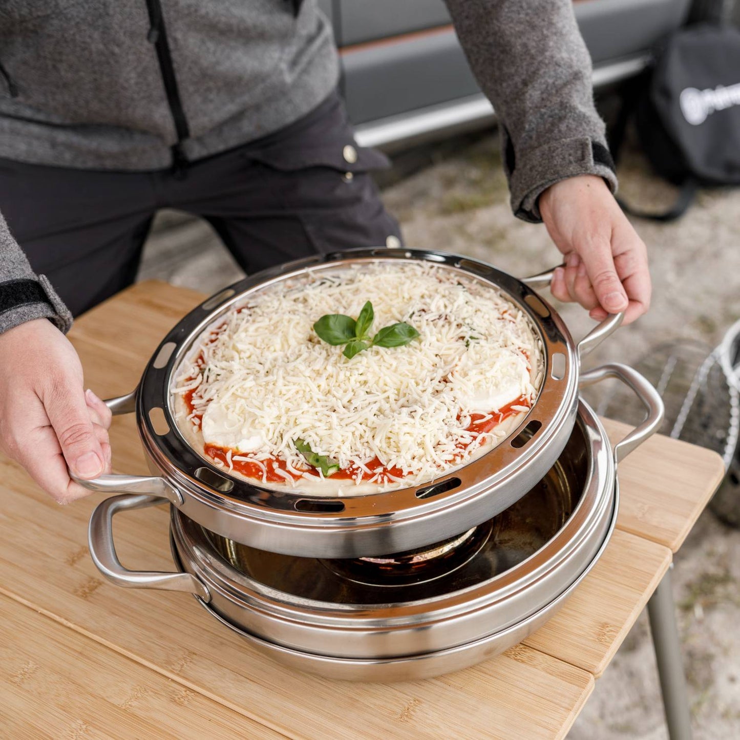 Round stainless steel pizza baking tray with handles and ventilation slots, holding an uncooked pizza topped with cheese and basil, designed for use in camping ovens, featured in Farm, Garden & Tool, Camping, Best Sellers, and Promo Eligible collections.