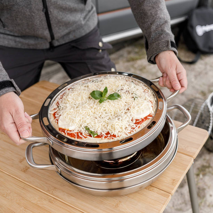 Round stainless steel pizza baking tray with handles and ventilation slots, holding an uncooked pizza topped with cheese and basil, designed for use in camping ovens, featured in Farm, Garden & Tool, Camping, Best Sellers, and Promo Eligible collections.