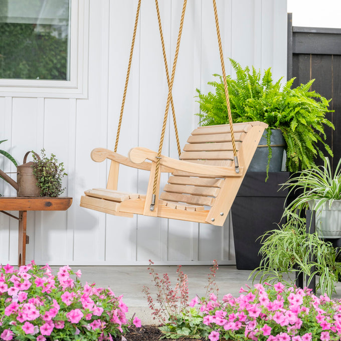 Handcrafted cypress porch swing with smooth wooden slats and sturdy rope suspension, set on a porch surrounded by vibrant pink flowers and lush green plants in outdoor home decor collections.