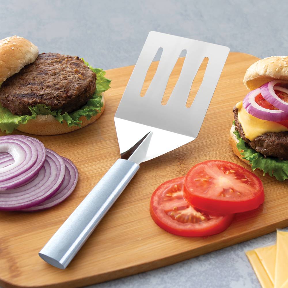Sturdy stainless steel spatula with a slotted, angled face and a durable handle, shown with fresh burger ingredients on a wooden cutting board, part of Kitchen & Food, Storage and Serving, USA Made, Best Sellers, and Promo Eligible collections.