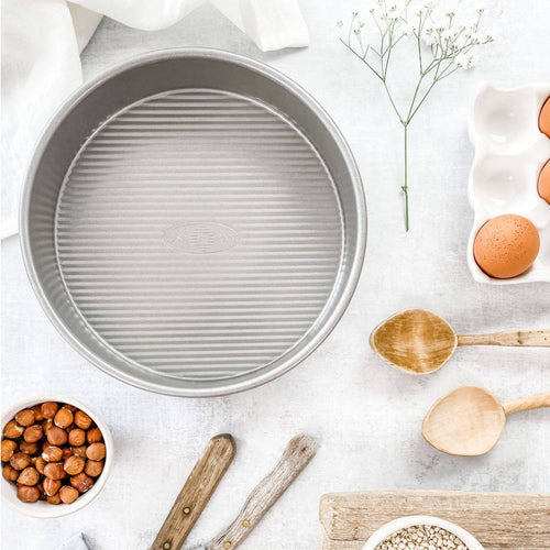Round aluminized steel cake pan with a fluted surface for even baking and easy release, surrounded by wooden spoons, eggs, and nuts, ideal for Kitchen & Food and USA Made collections.