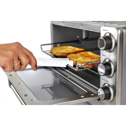 Hand gripping a white toaster tong with serrated teeth lifting toast from a metal oven rack in a compact stainless steel toaster oven, part of Kitchen Gadgets and Cooking and Baking Helpers collections.