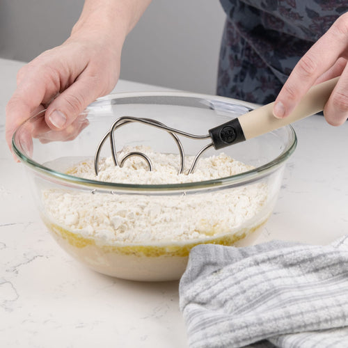 Handheld dough whisk with a sturdy full tang design mixing flour and liquid in a glass bowl, shown in a modern kitchen setting with a gray striped towel nearby, ideal for Baking Supplies and Kitchen Gadgets collections.