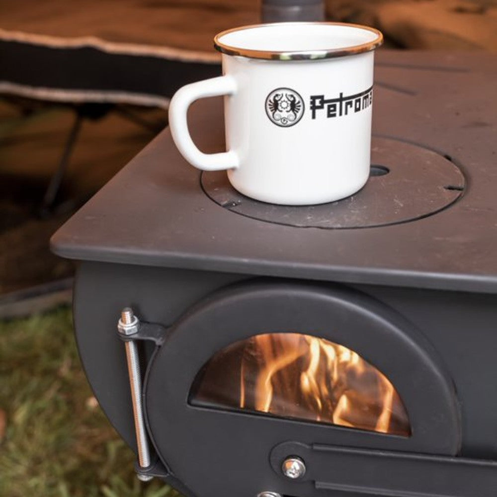 White enameled steel mug with metal rim and black logo sitting on a wood stove with visible flames inside, perfect for Farm, Garden & Tool, Camping, Brewing Coffee and Tea, Kitchen & Food, Dinnerware, Best Sellers, and Promo Eligible collections.