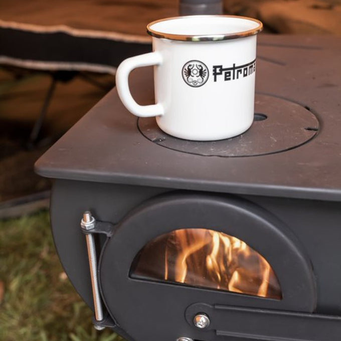 White enameled steel mug with metal rim and black logo sitting on a wood stove with visible flames inside, perfect for Farm, Garden & Tool, Camping, Brewing Coffee and Tea, Kitchen & Food, Dinnerware, Best Sellers, and Promo Eligible collections.