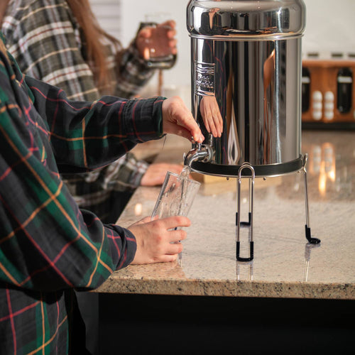 Person filling a clear glass with filtered water from a polished stainless steel gravity water filter elevated on a sturdy stand in a kitchen setting, featured in Appliances and Emergency Supplies collections.
