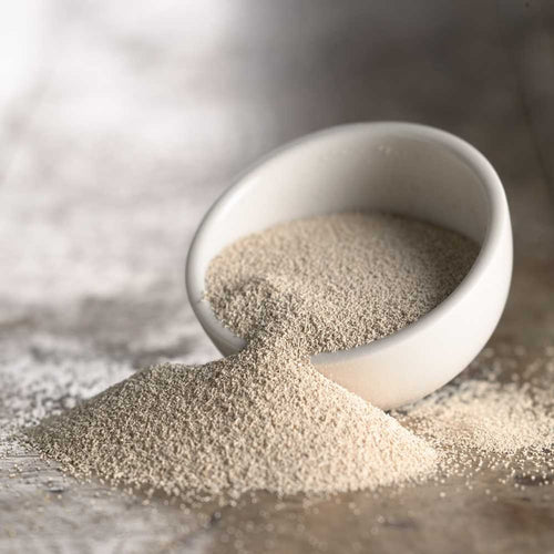 White ceramic bowl tipped over with fine beige active dry yeast granules spilling onto a textured surface, featured in Kitchen & Food, Dry Goods and Mixes, and Bread Making Supplies collections.