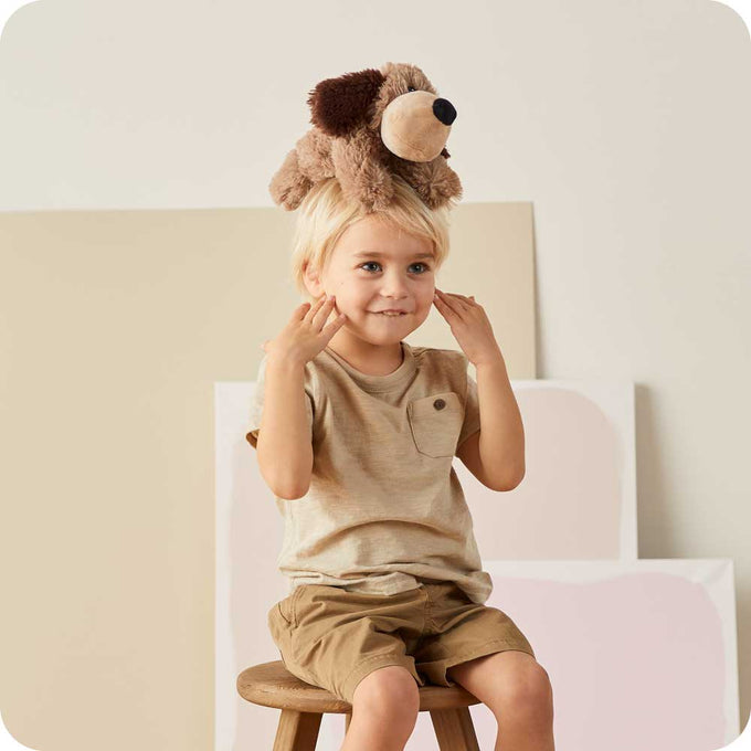 Young child sitting on a wooden stool wearing a soft brown plush dog toy on their head, perfect for Health & Wellness and Children's Toys collections.