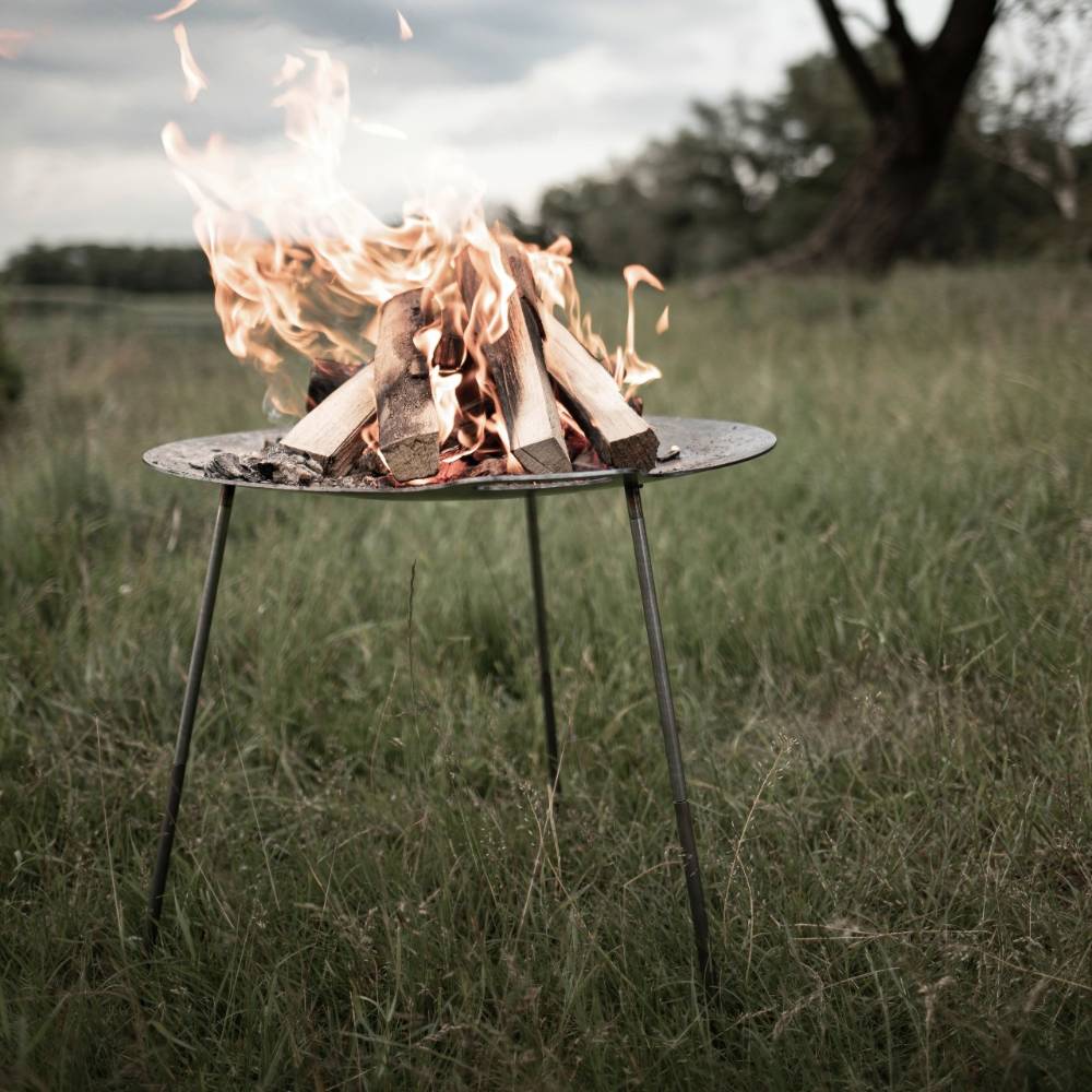 Steel fire bowl with three slender legs holding burning wood, creating a warm and inviting campfire atmosphere in a grassy outdoor setting, perfect for Farm, Garden & Tool, Camping, Best Sellers, and Promo Eligible collections.