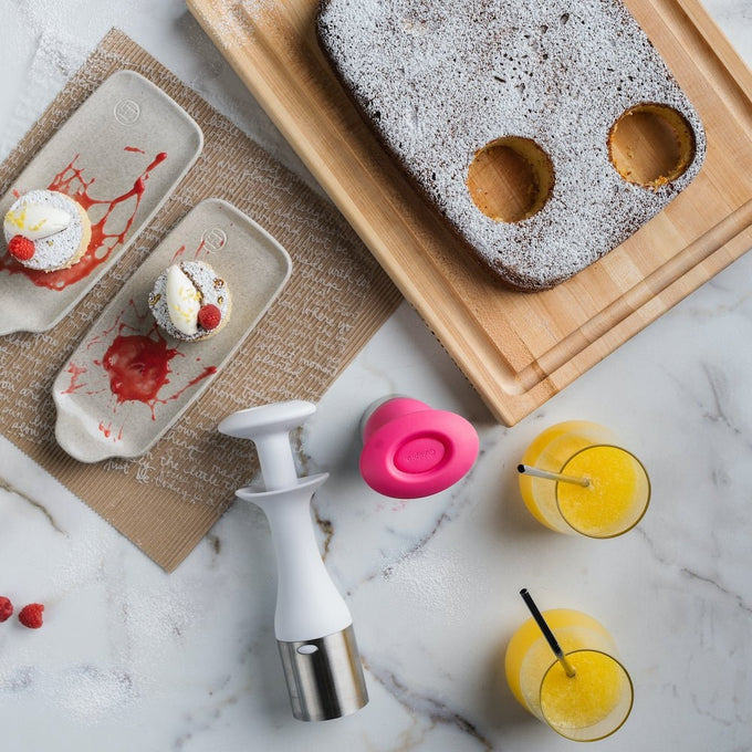 Ergonomic ice cream scoop with button release beside a powdered cake with scooped circles, two dessert plates with fruit-topped treats, and glasses of juice on a kitchen surface.