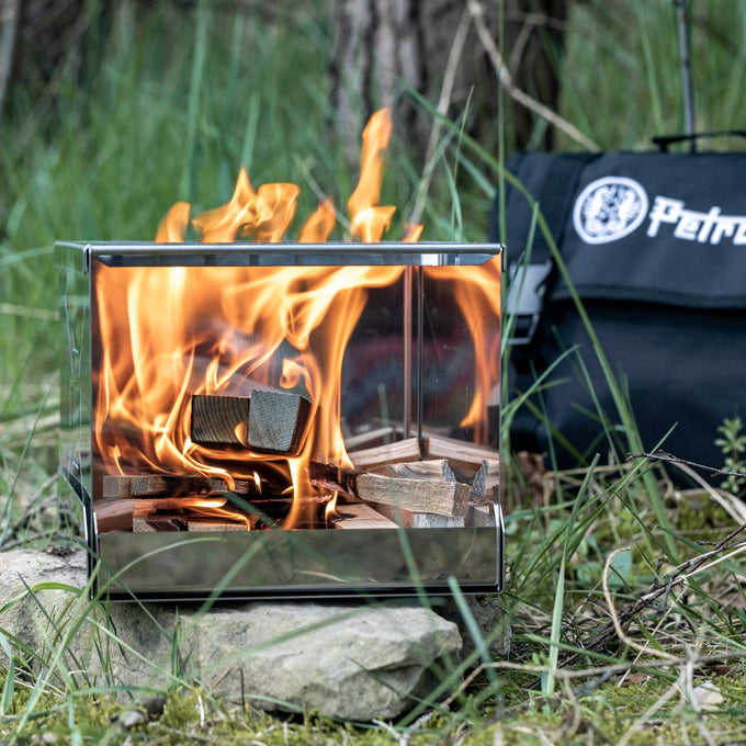 Handcrafted compact stainless steel camp stove actively burning wood, displayed outdoors on natural stone with branded carrying case, perfect for Farm, Garden & Tool, Camping, Emergency Supplies, Best Sellers, and Promo Eligible collections.
