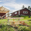 Portable chain sawmill set up outdoors next to a wooden framework under construction near a red farmhouse, highlighting wood cutting and hauling in farm, garden, and tool settings.