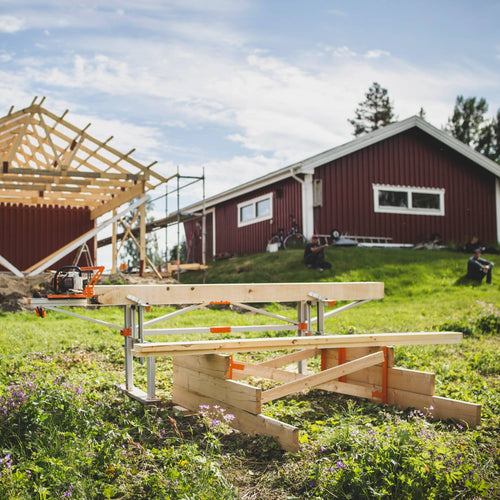 Portable chain sawmill set up outdoors next to a wooden framework under construction near a red farmhouse, highlighting wood cutting and hauling in farm, garden, and tool settings.